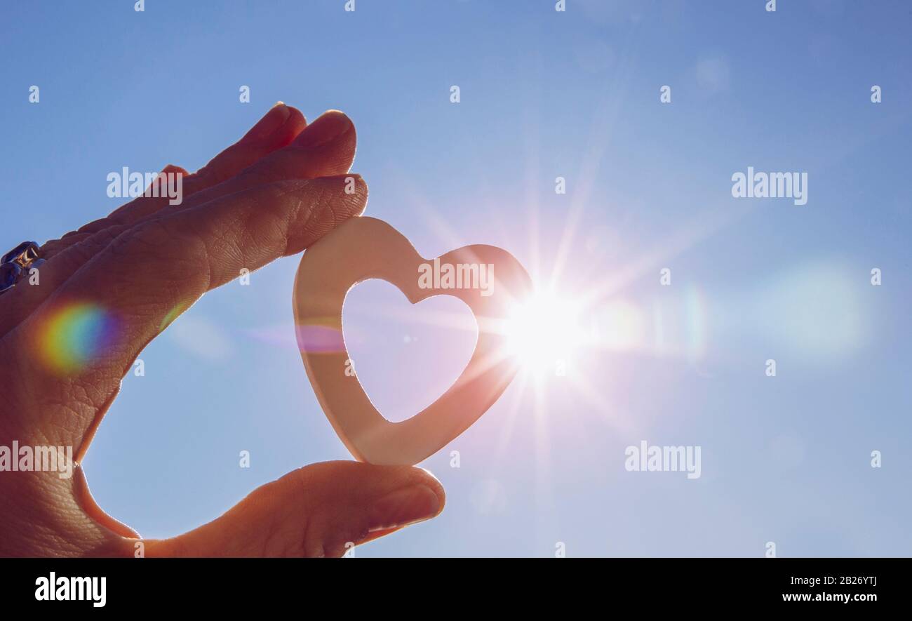 Close up view of woman hand holding white wooden heart against sun and blue sky with lens flare and sun`s photography star effect. Positive emotions. Stock Photo